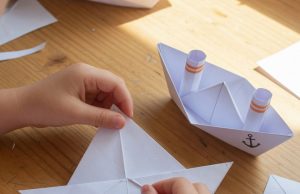 Comment fabriquer un bateau en origami : un cours de maître classique pour les enfants et les parents.⛵ L'enfant plie un bateau en origami à partir de papier, un exemplaire fini est posé sur la table.